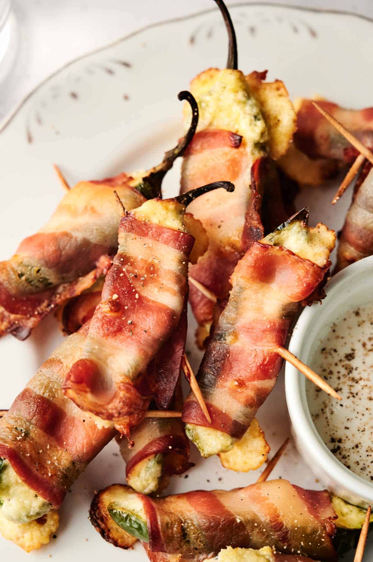 Bacon-wrapped jalape&ntilde;o poppers with a creamy cheese filling sit enticingly on a plate, each secured with toothpicks. A small bowl of dipping sauce accompanies these delicious appetizers.