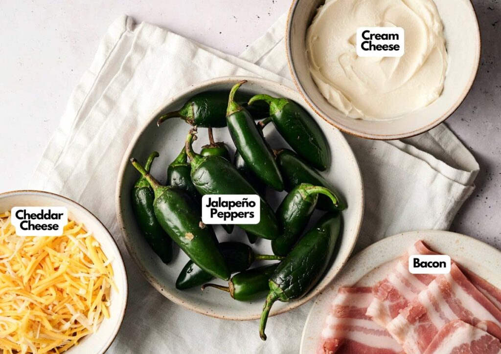 A bowl of jalapeño peppers, a bowl of shredded cheddar cheese, a bowl of cream cheese, and a plate of raw bacon on a white cloth—perfect ingredients for making delicious bacon wrapped jalapeno poppers.