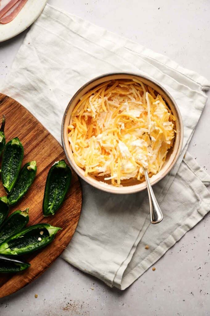 A bowl of shredded cheese and cream cheese with a spoon sits next to halved jalapeños on a wooden cutting board, ready to become delicious bacon-wrapped jalapeño poppers.
