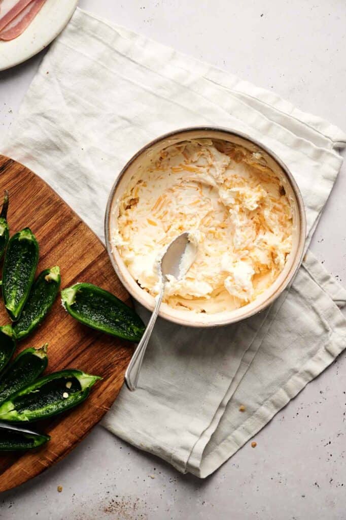 A bowl of cheese mixture with a spoon resting inside sits on a cloth next to a wooden board featuring sliced green peppers, evoking the start of a mouthwatering batch of bacon-wrapped jalapeno poppers.