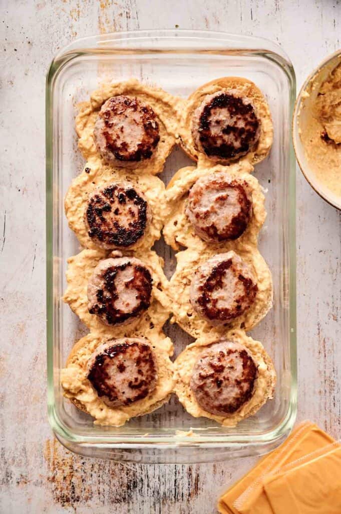 A glass baking dish holds eight mouthwatering sausage patties, each resting on a bed of creamy hummus, reminiscent of the flavor-packed indulgence found in cheeseburger sliders. Nearby, there's a bowl with extra hummus for added delight.