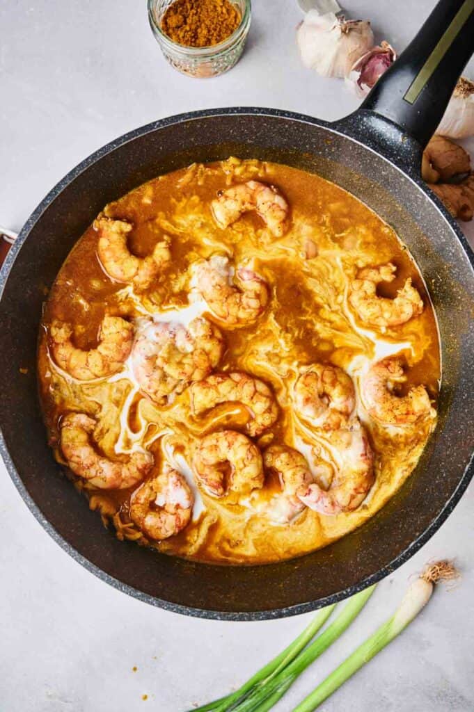 A pot filled with shrimp cooking in a rich, golden Thai coconut curry sauce, surrounded by garlic cloves, a jar of spices, and green onions on a light-colored surface.