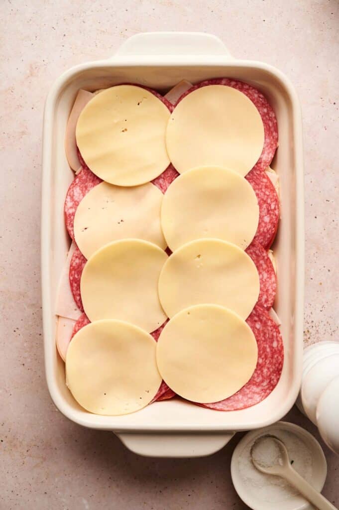 A casserole dish filled with layers of sliced salami and round provolone cheese slices, reminiscent of Italian Sandwich Sliders.