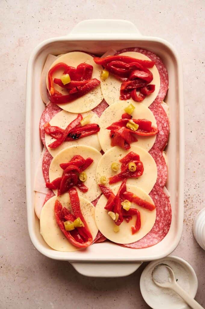 A white baking dish brimming with layers of salami, provolone cheese, red peppers, and sliced green olives offers the perfect foundation for delicious Italian Sandwich Sliders.