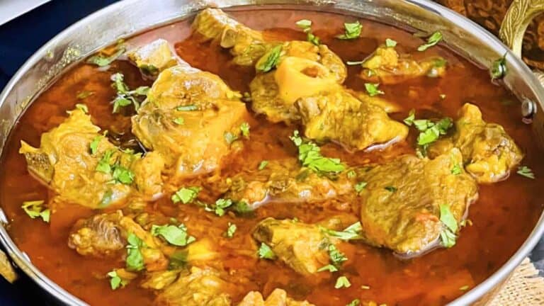 A metal bowl filled with mutton curry, garnished with chopped cilantro. The rich, red sauce surrounds pieces of meat.