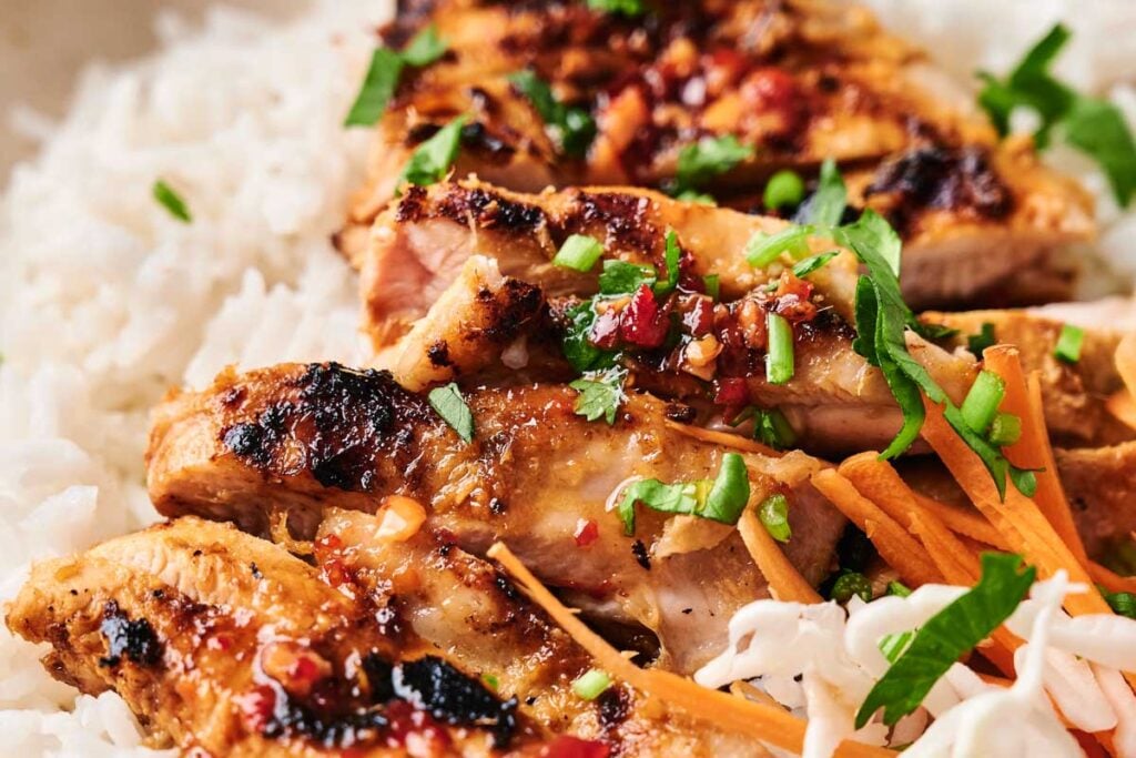 Lemongrass chicken slices topped with sauce and chopped herbs, served on white rice with shredded carrots and cabbage.