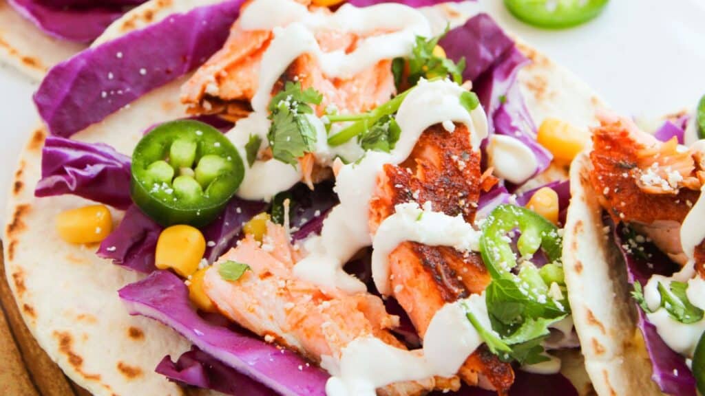 Close-up of tacos with grilled salmon, purple cabbage, corn, jalape&ntilde;o slices, cilantro, and a drizzle of white sauce on flour tortillas.