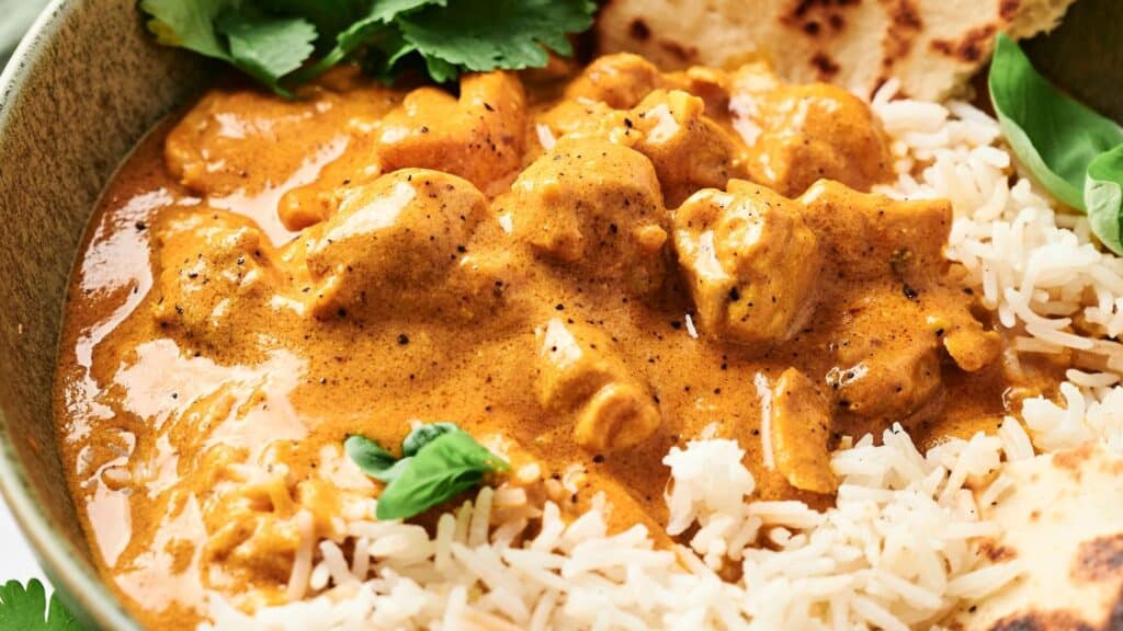 A bowl of creamy orange curry with chunks of meat, served with white rice and garnished with cilantro, alongside a piece of naan bread.