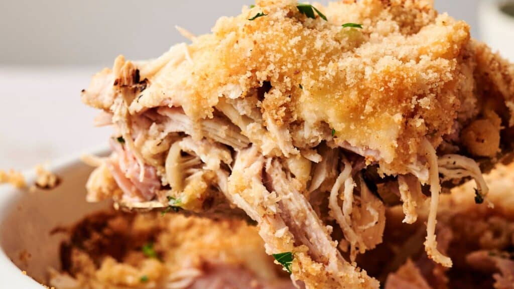 Close-up of a serving of shredded chicken topped with breadcrumbs and herbs, being lifted from a baking dish.