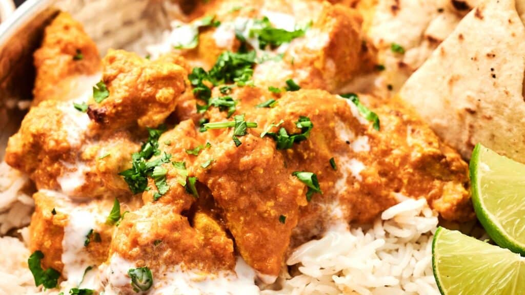 A close-up of chicken tikka masala with rice, garnished with fresh cilantro. Lime wedges and naan are on the side.