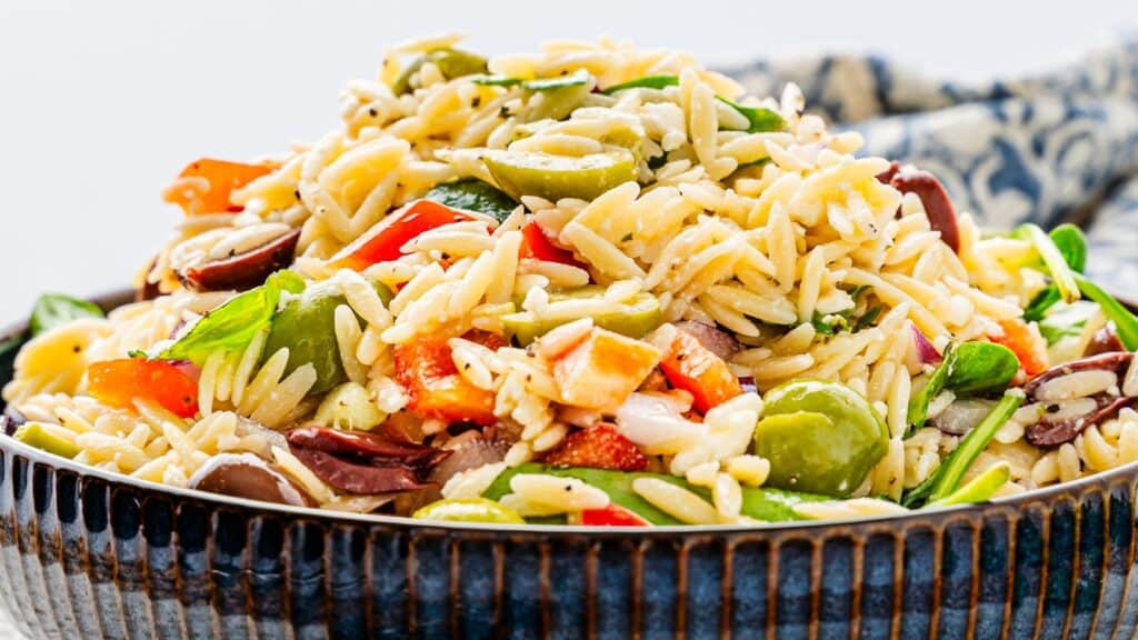 A bowl of orzo pasta salad with olives, bell peppers, and leafy greens.
