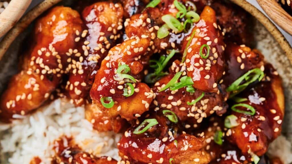Close-up of a dish featuring glazed chicken pieces topped with sesame seeds and green onions, served over white rice.