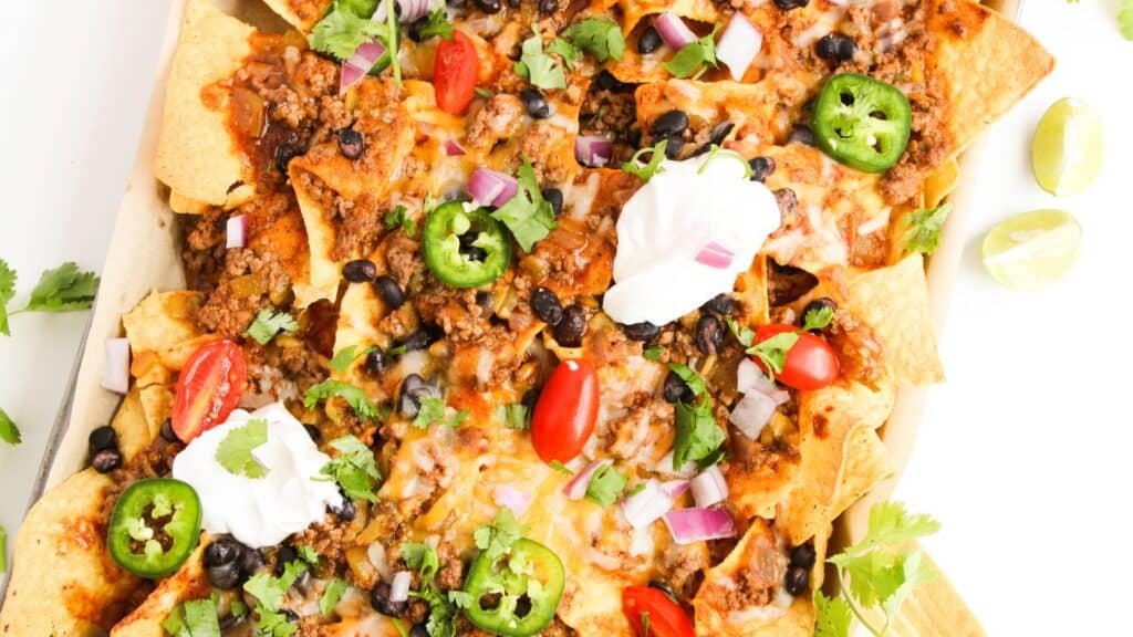A tray of loaded nachos topped with melted cheese, ground beef, jalapeño slices, cherry tomatoes, black beans, onions, cilantro, and dollops of sour cream, with lime wedges on the side.