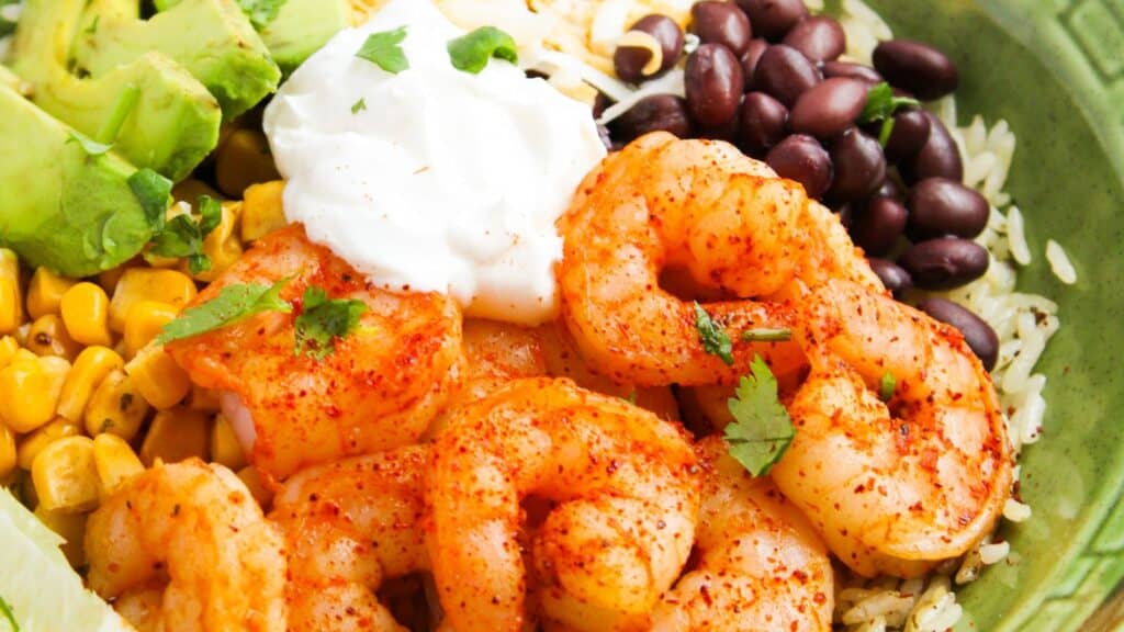 A bowl with seasoned shrimp, avocado, black beans, corn, rice, shredded cheese, and a dollop of sour cream, garnished with cilantro.