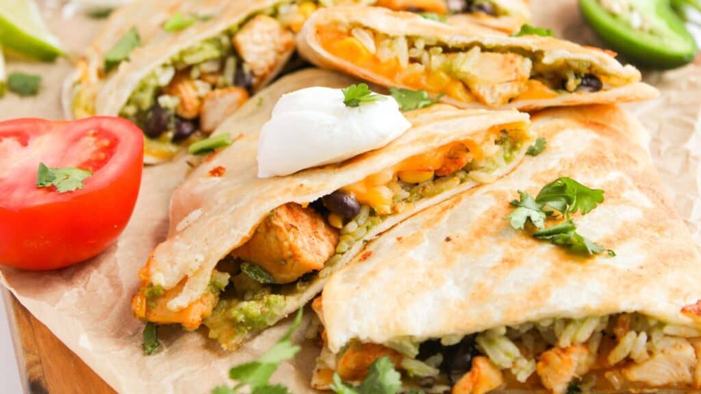 Close-up of sliced quesadillas filled with chicken, black beans, rice, and cheese, topped with sour cream and cilantro. Sliced tomato and jalapeño on the side.