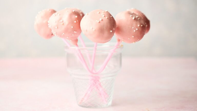 Pink cake pops with white sprinkles on sticks in a clear glass jar against a light background.