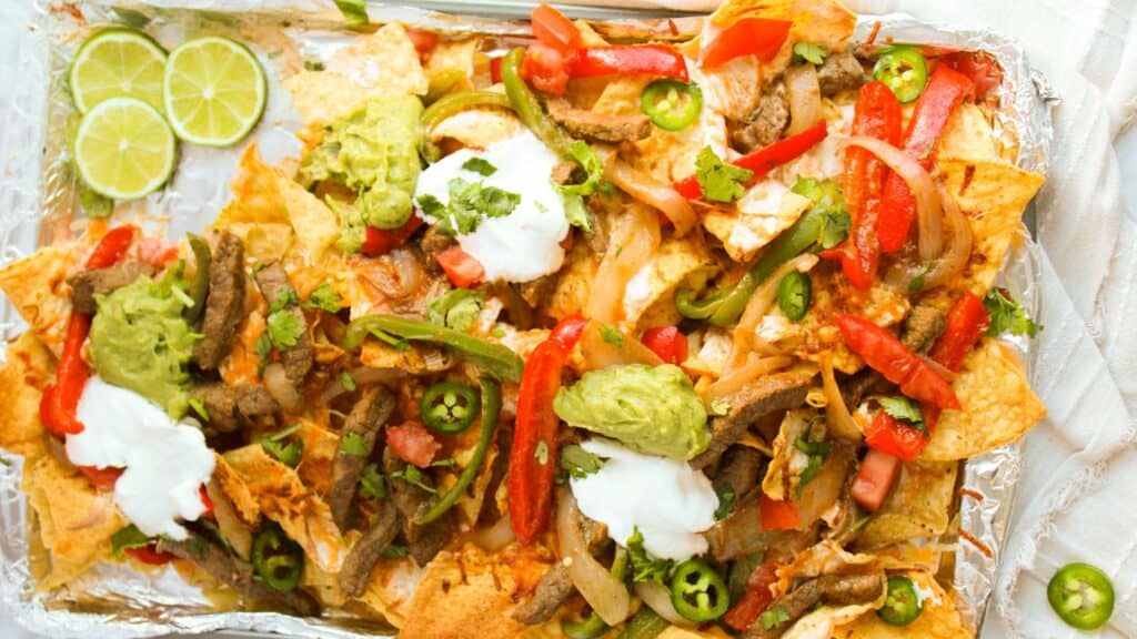 A tray of loaded nachos topped with grilled meat, sliced peppers, jalape&ntilde;os, guacamole, sour cream, and cilantro, garnished with lime slices.