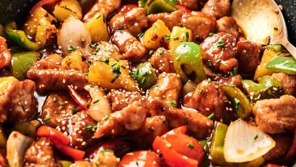 Close-up of a colorful sweet and sour pork dish with chunks of pork, bell peppers, onions, and pineapple, garnished with sesame seeds and chives.