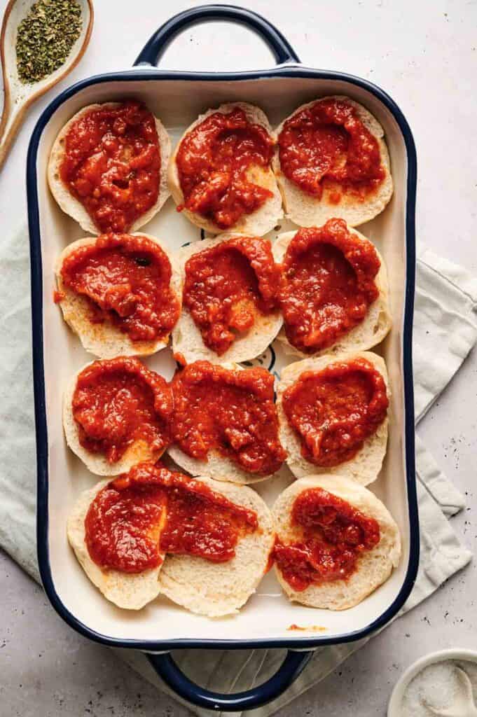 A baking dish with sliced bread rolls, transformed into delicious meatball sliders, is spread with tomato sauce and arranged neatly on a white surface.