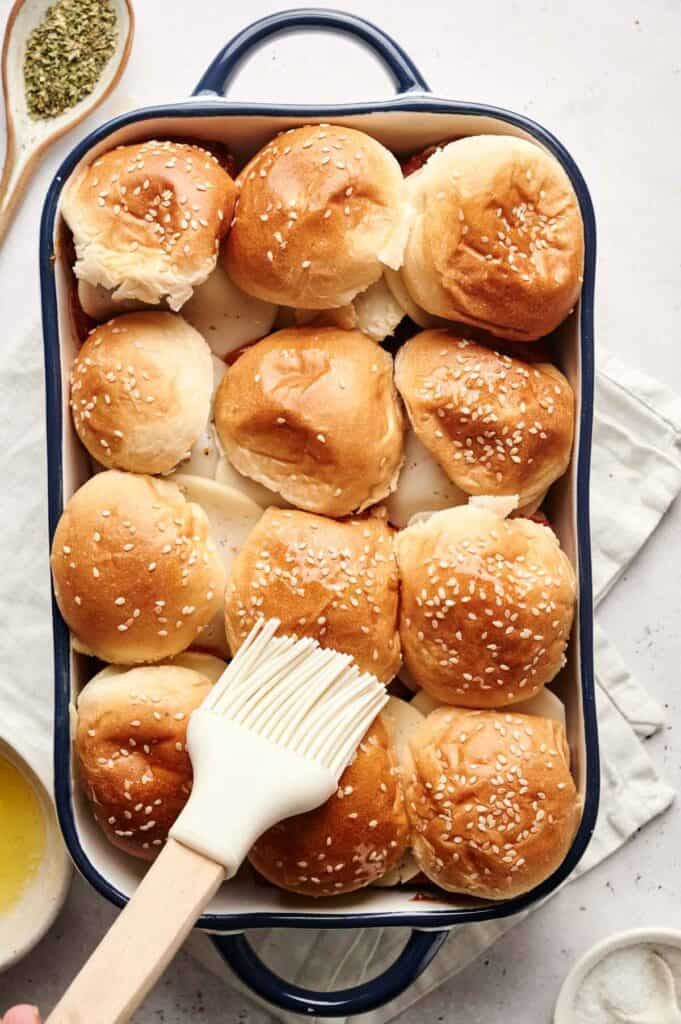 A baking dish filled with freshly baked dinner rolls topped with sesame seeds. Perfect as a base for meatball sliders, they are being coated with melted butter using a brush, enhancing their golden crust.