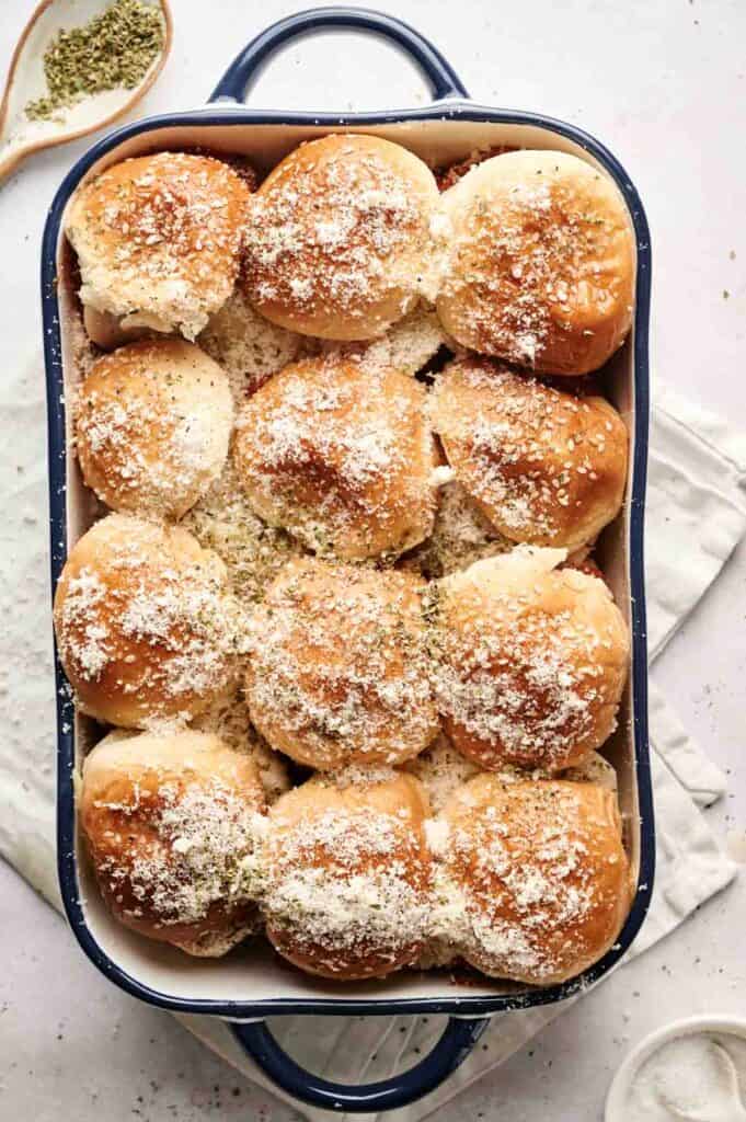 A baking dish filled with golden-brown meatball sliders topped with grated cheese and herbs.
