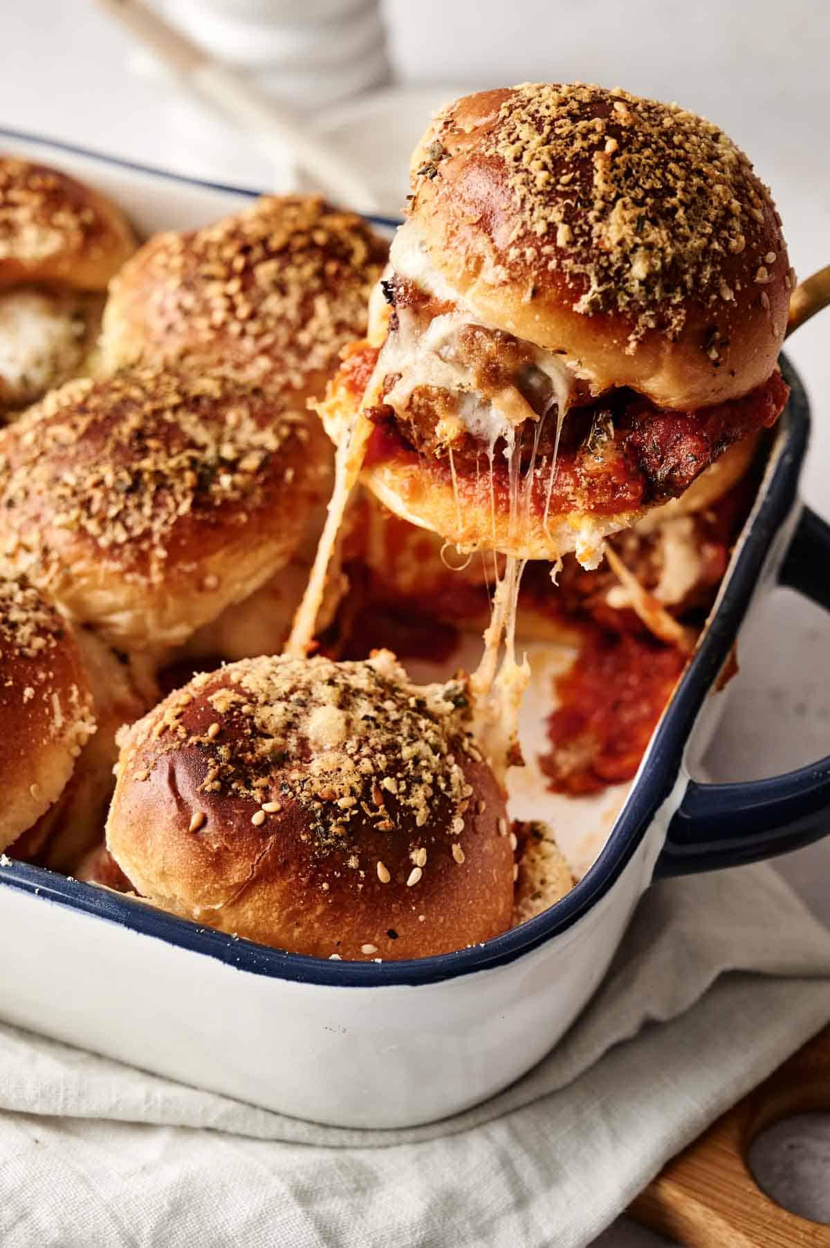 A dish of delicious meatball sliders with melted cheese is presented, with one slider being lifted to reveal its cheesy texture. The crispy-topped sliders are nestled in a rectangular pan, perfectly capturing the essence of comfort food.
