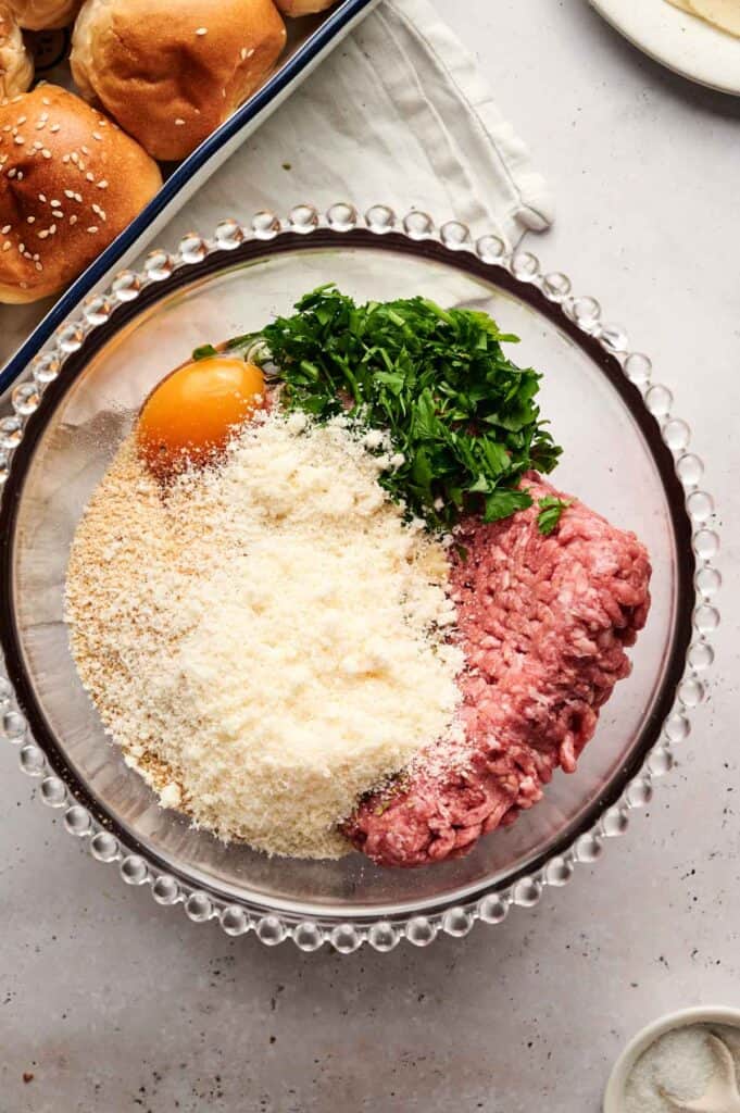 A glass bowl with ground meat, breadcrumbs, egg, chopped parsley, and grated cheese sits ready for mixing&mdash;a perfect start for delicious meatball sliders.