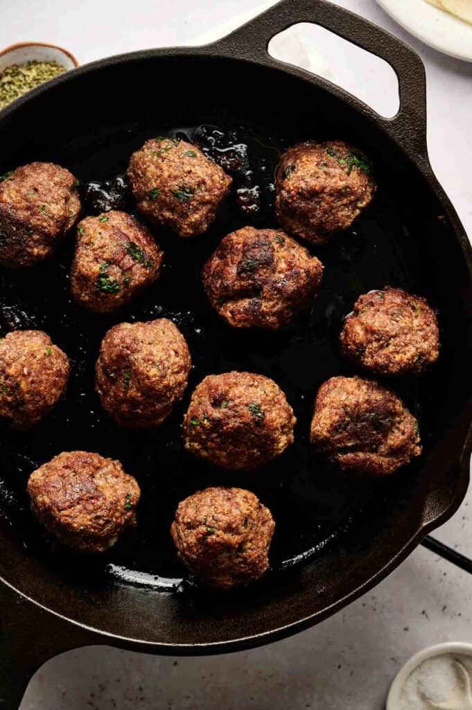 A black skillet with twelve browned meatballs cooking, some herbs visible in the mixture, perfect for crafting delicious meatball sliders.