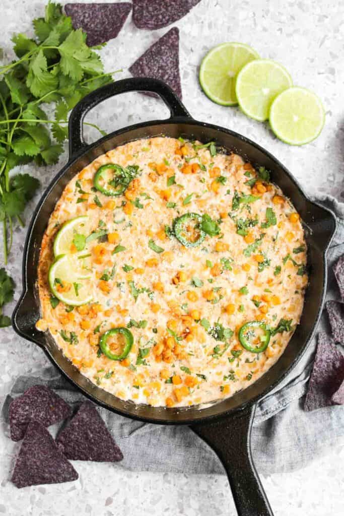 A cast iron skillet brimming with Mexican Street Corn Dip, topped with sliced jalapeños, lime wedges, and cilantro. Blue corn tortilla chips are scattered around for a perfect crunchy finish.