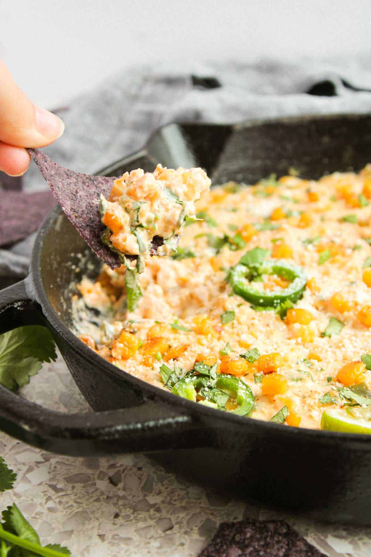 A hand holding a tortilla chip scoops up a creamy Mexican Street Corn Dip, beautifully garnished with sliced jalape&ntilde;os and fresh herbs, all served in a rustic cast iron skillet.