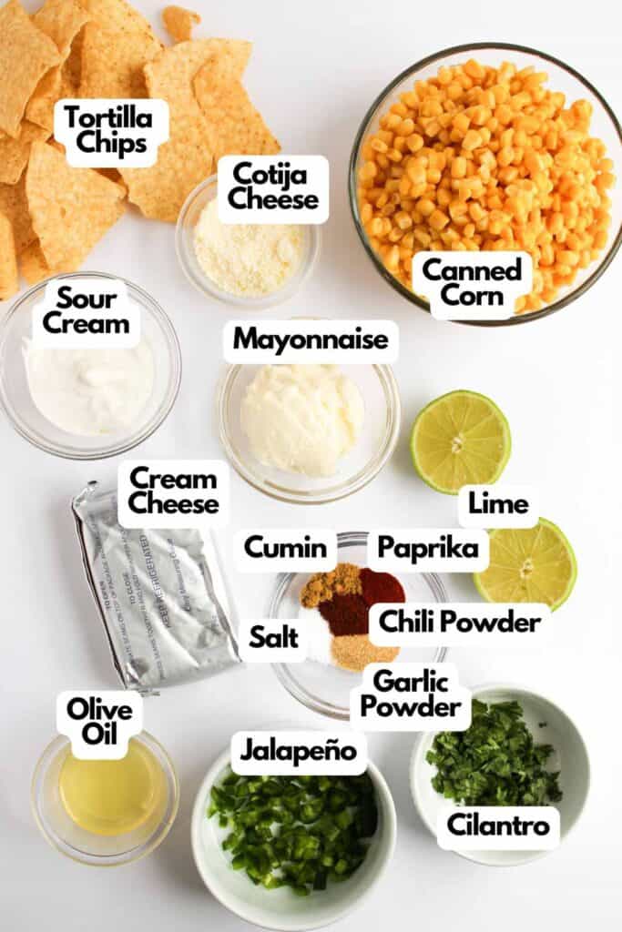 The ingredients for Mexican Street Corn Dip are artfully arranged on a white surface, showcasing tortilla chips, canned corn, sour cream, mayonnaise, and cream cheese. Adding cotija cheese, lime, spices, olive oil, jalape&ntilde;o, and cilantro brings a vibrant splash of flavor to this dish.