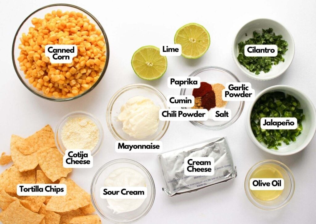 Ingredients for a Mexican Street Corn Dip recipe are arranged on a white surface, including canned corn, lime, cilantro, paprika, cumin, chili powder, salt, garlic powder, jalapeño, olive oil, cream cheese, mayo, sour cream, cotija cheese, and tortilla chips.