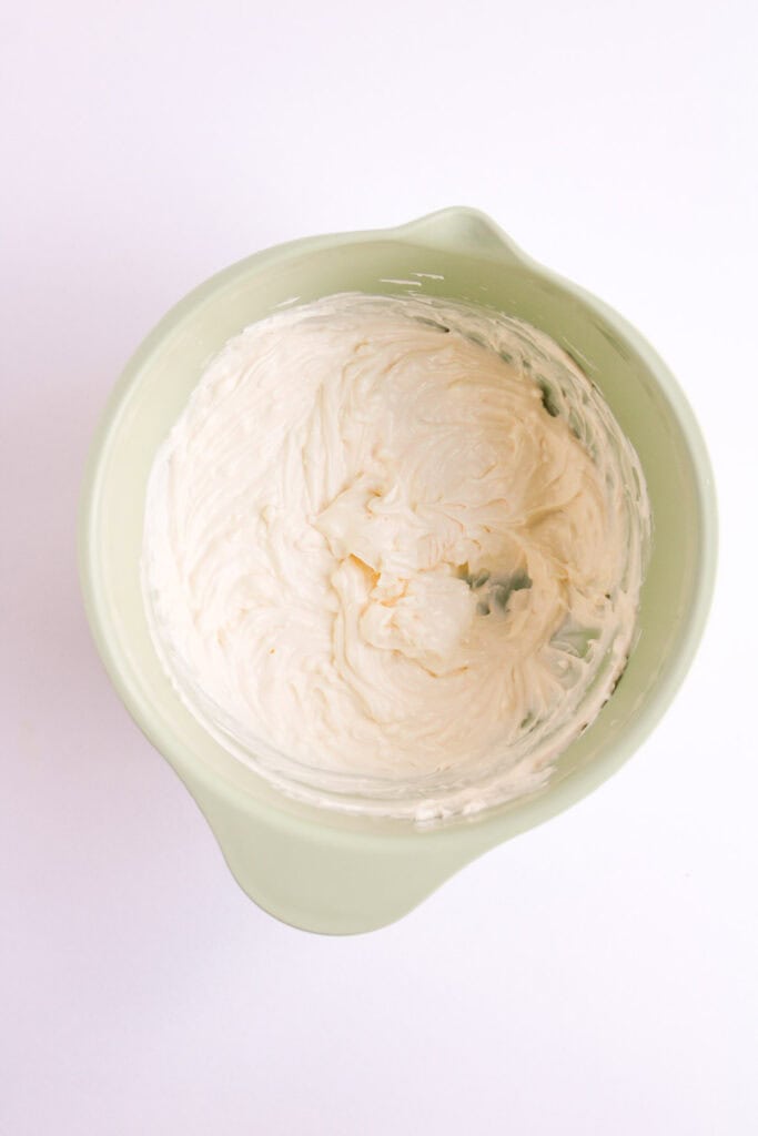 In a green bowl on a white background, the creamy, smooth white frosting evokes the vibrant textures of a Mexican Street Corn Dip.