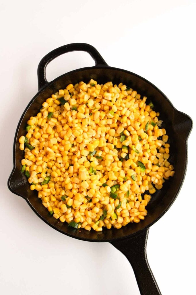 Cast iron skillet filled with cooked yellow corn kernels and jalapeños on a white background, reminiscent of a vibrant Mexican Street Corn Dip.