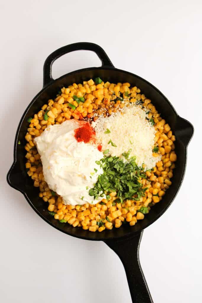 A black skillet brimming with Mexican Street Corn Dip: corn topped with sour cream, cheese, cilantro, and a dash of paprika, set against a crisp white background.