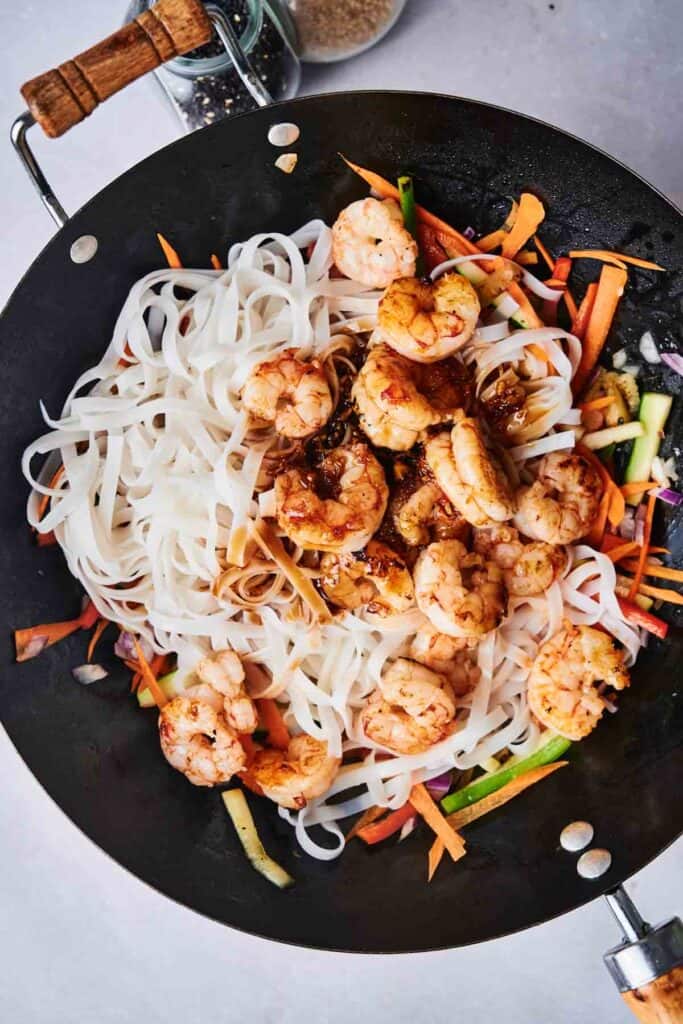 A wok filled with Thai drunken noodles featuring cooked shrimp, rice noodles, and sliced vegetables like carrots and zucchini rests on a light gray surface.