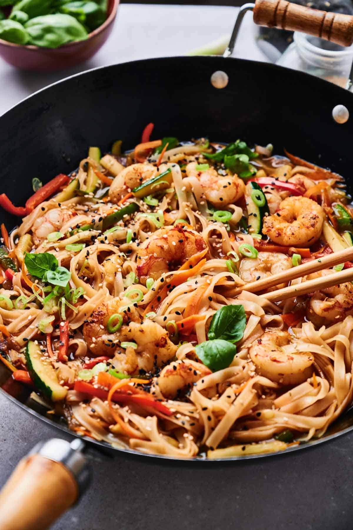 A wok filled with Thai drunken noodles, shrimp, and vegetables, garnished with sesame seeds, green onions, and basil leaves. Chopsticks rest on the side.
