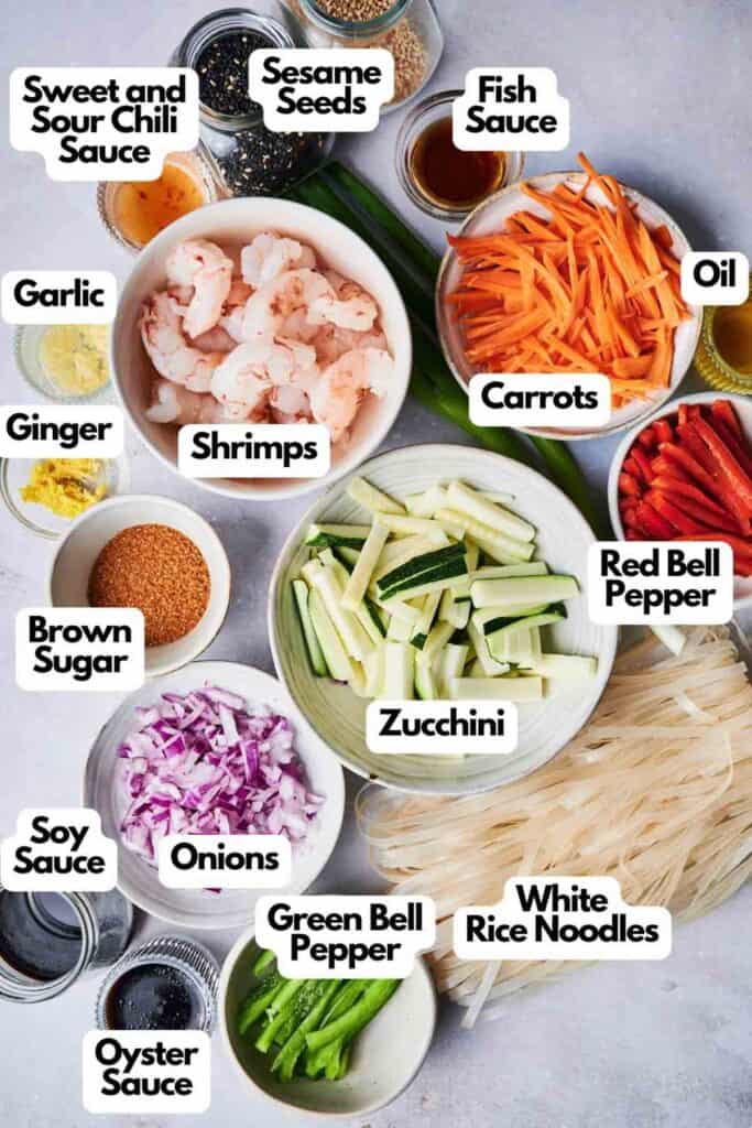 Ingredients for Thai drunken noodles laid out, featuring shrimps, zucchini, onions, carrots, bell peppers, noodles, garlic, ginger, and assorted sauces and seeds—all neatly labeled for easy identification.