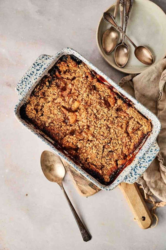 A baked apple crumble in a speckled rectangular dish on a wooden board, accompanied by a serving spoon and a stack of plates with spoons on the side.