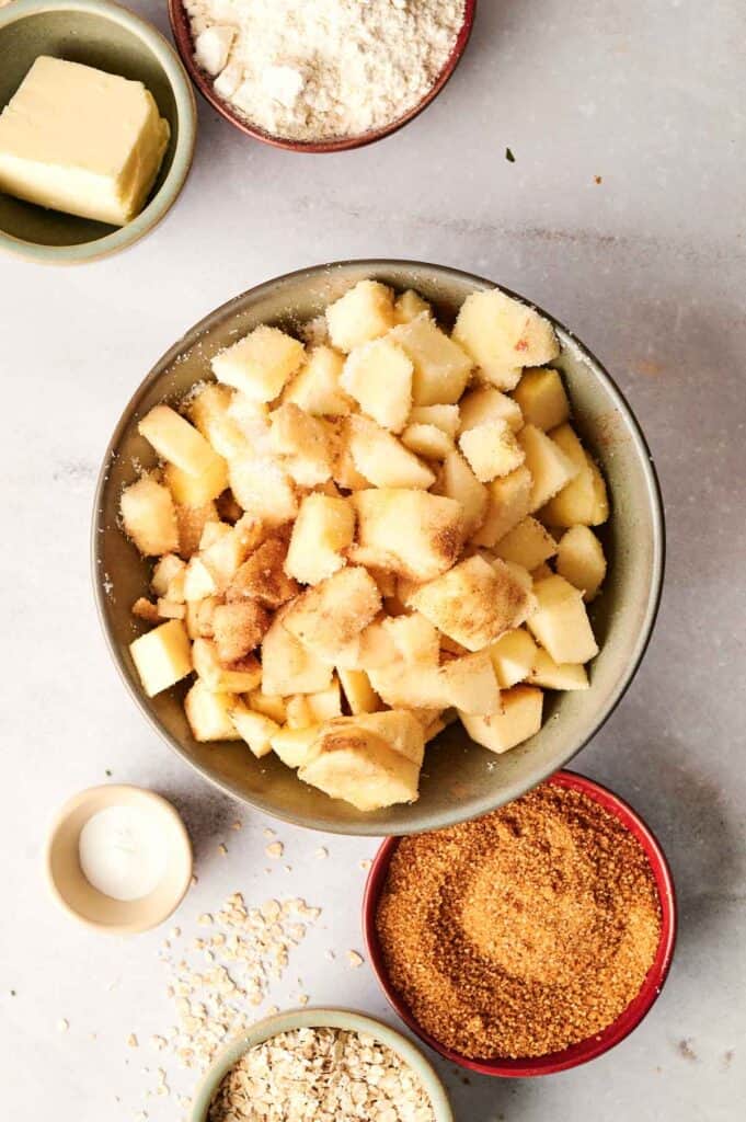 Bowl of chopped apples with sugar and cinnamon, surrounded by small bowls of oats, brown sugar, flour, and a stick of butter on a gray surface.