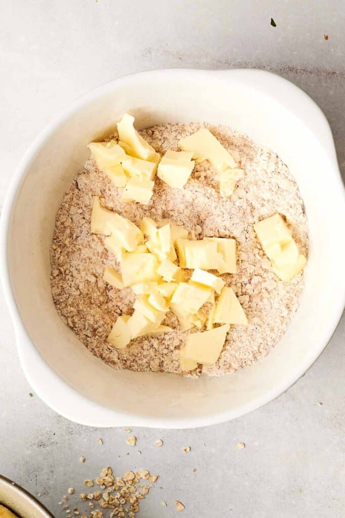 A white bowl contains flour and oats with butter chunks on top. Some oats are scattered on the table.
