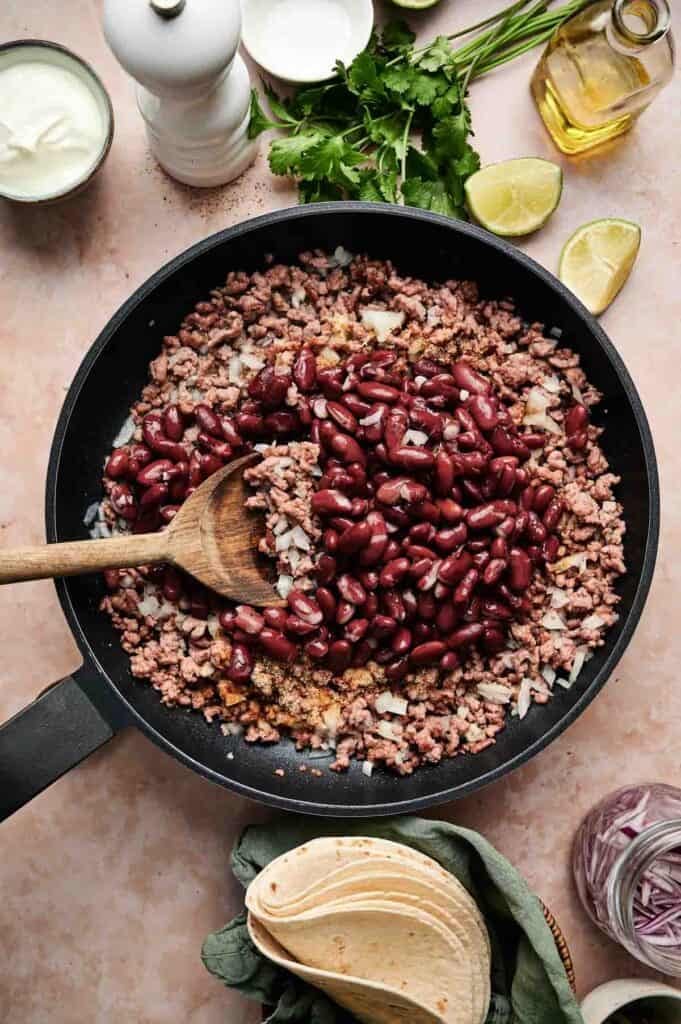 A skillet with cooked ground beef enchilada casserole, kidney beans, and onions is stirred with a wooden spoon. Nearby are tortillas, sliced onions, lemon wedges, and herbs.