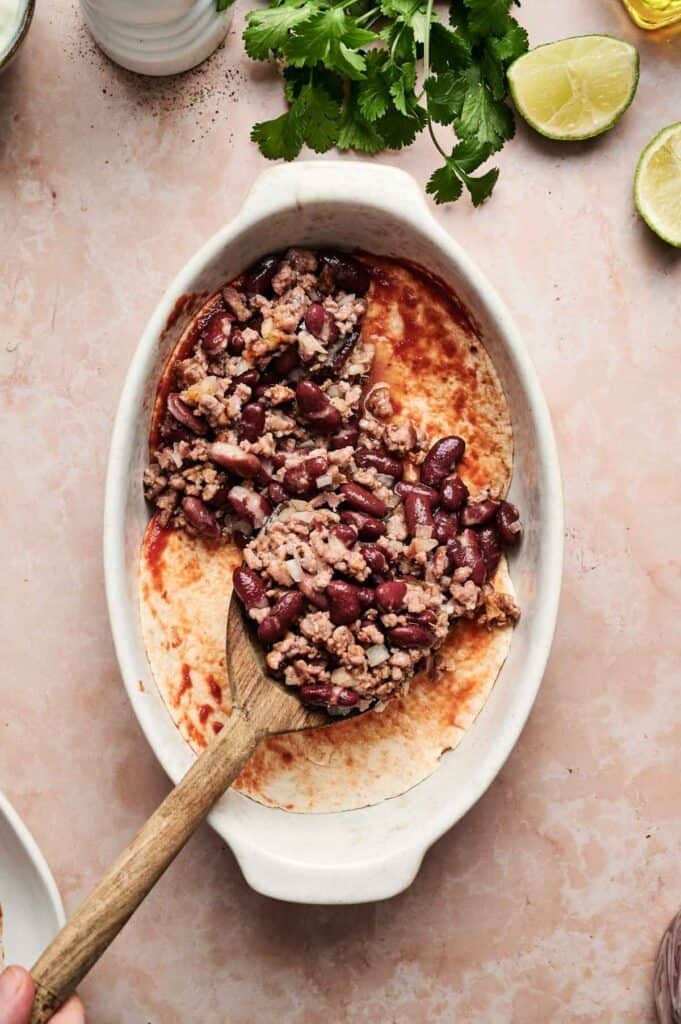A wooden spoon rests in an oval dish containing cooked minced meat and kidney beans, reminiscent of a hearty beef enchilada casserole. Fresh cilantro and lime are nearby on the marble surface.