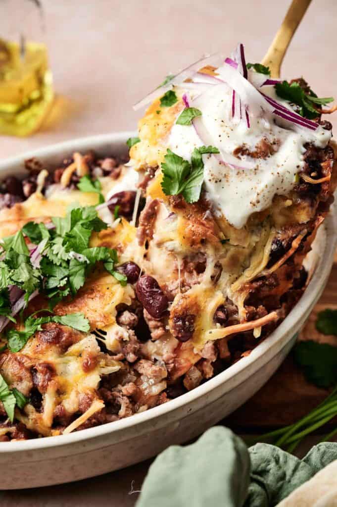 A close-up of a cheesy beef enchilada casserole with ground meat and beans, topped with sour cream and garnished with cilantro and sliced onions. A spoon lifts a portion from the baking dish.