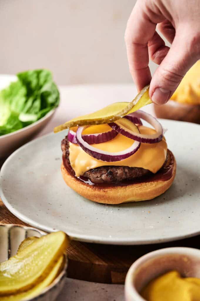 A hand carefully places a pickle slice on a mouthwatering cheeseburger, with crispy onion rings and fresh lettuce adding to the delicious backdrop.