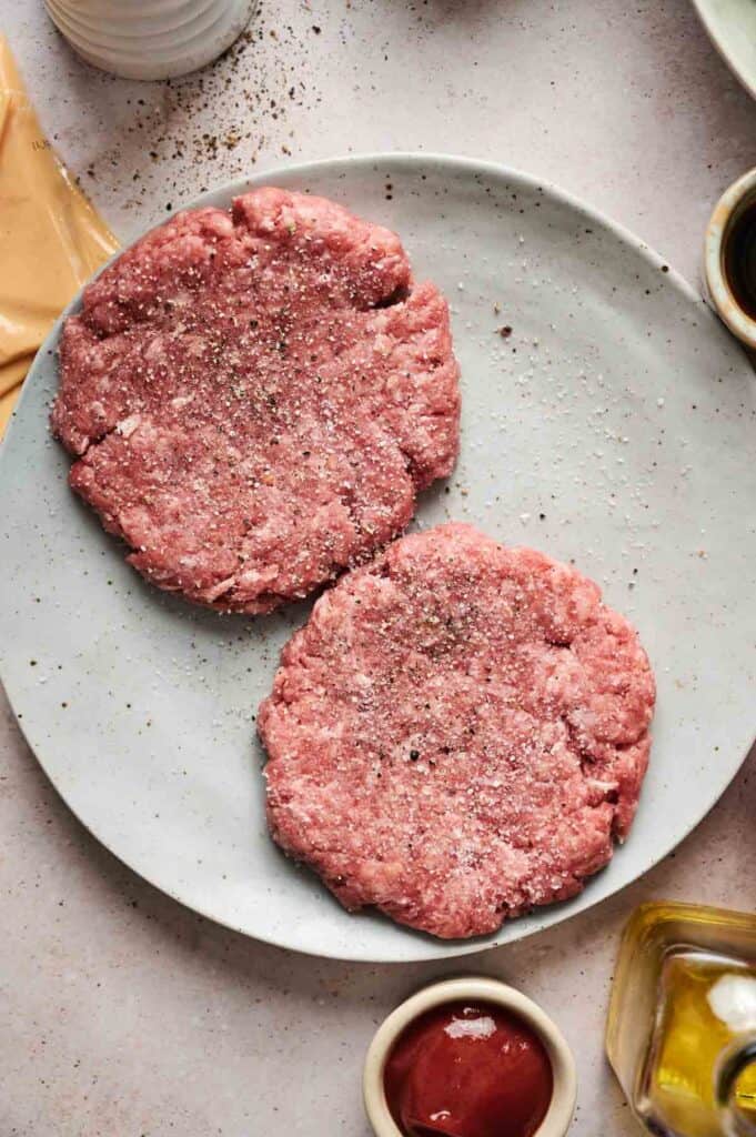 Two raw ground beef patties, seasoned with salt and pepper, await their transformation into a delicious cheeseburger on a round plate, surrounded by small bowls and ingredients on a light surface.