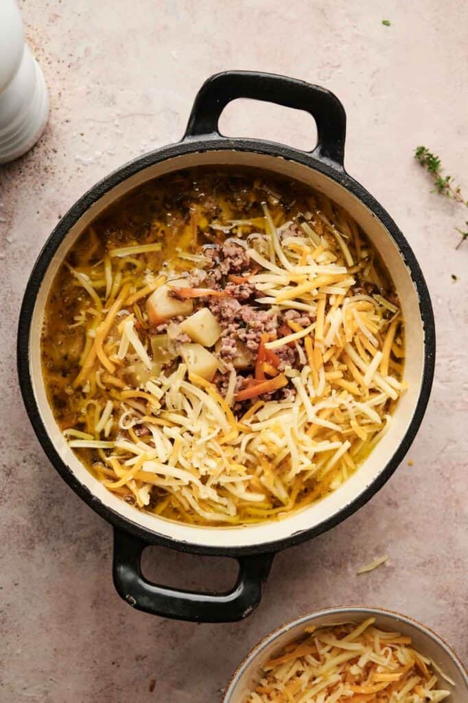 A pot of cheeseburger soup brimming with shredded cheese, cooked ground meat, diced potatoes, and chopped vegetables. The pot features two handles and rests on a light-colored surface.
