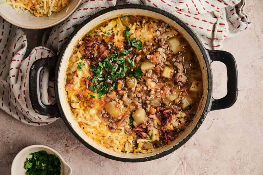 A simmering pot of stew reminiscent of a cheeseburger soup, featuring ground meat, potatoes, and savory herbs rests on a fabric surface. Nearby, a small bowl brims with freshly chopped parsley.