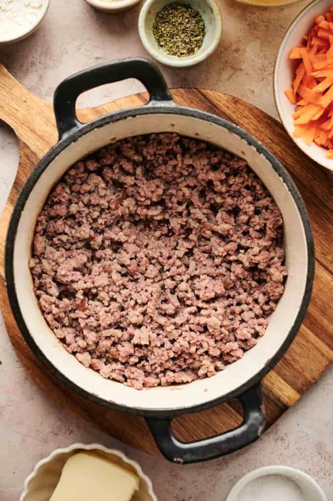 A pot filled with cooked ground meat sits on a wooden board, evoking the warm flavors of cheeseburger soup. It's surrounded by small bowls of sliced carrots, herbs, and butter, ready to enhance the comforting dish.