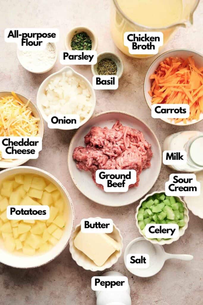 Ingredients for a cheeseburger soup recipe labeled clockwise from top: Chicken broth, carrots, milk, sour cream, celery, salt, pepper, butter, potatoes, ground beef, onion, cheddar cheese, flour, parsley, basil.